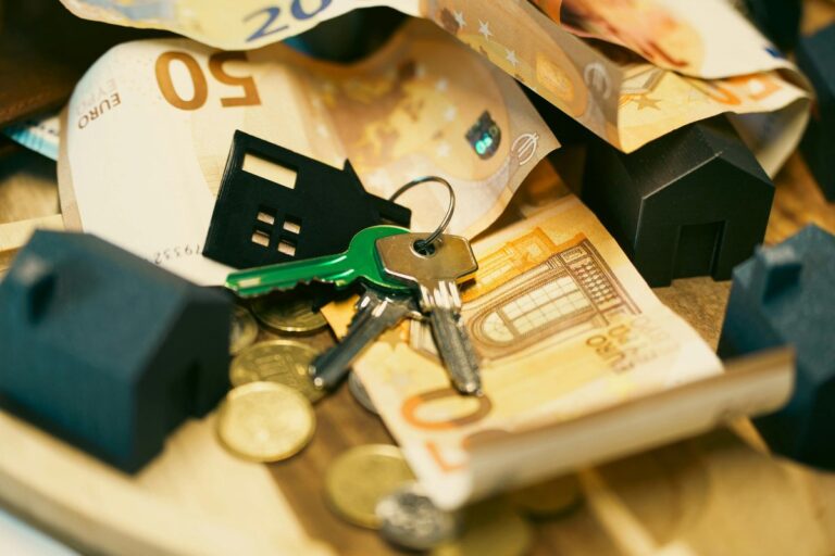 Klucze z brelokiem domku leżą na stosie banknotów euro i monet wśród miniaturowych domków
