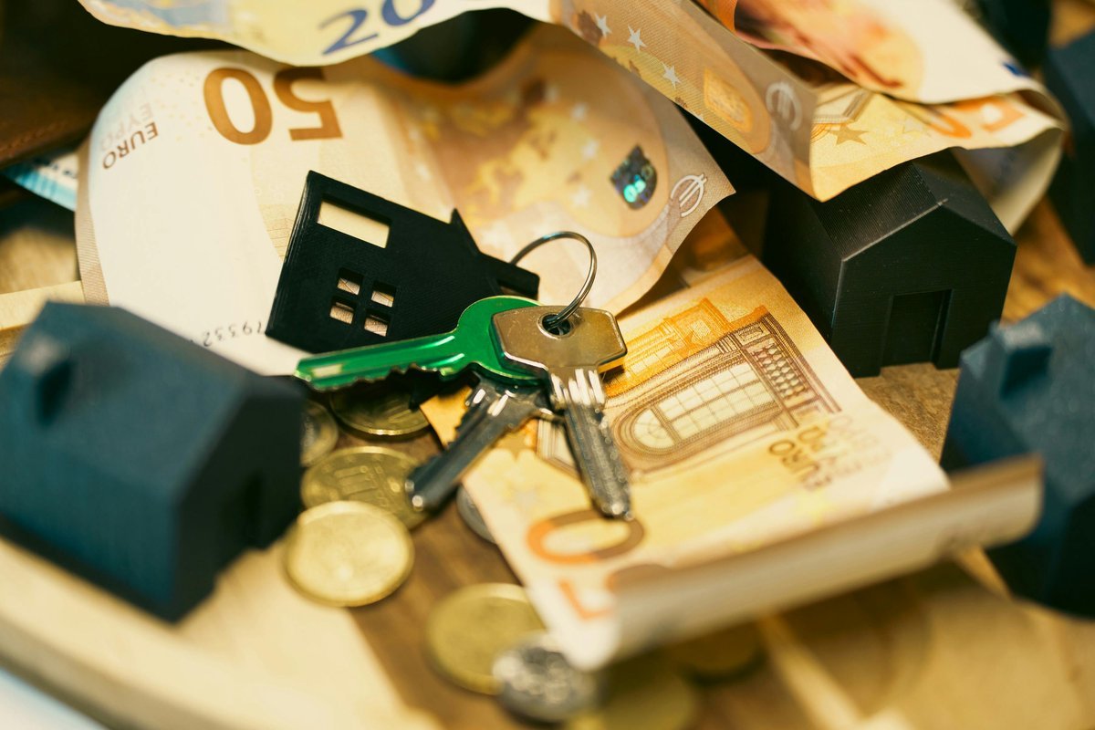 Keys with a house-shaped fob lying on a pile of euro banknotes and coins among miniature houses