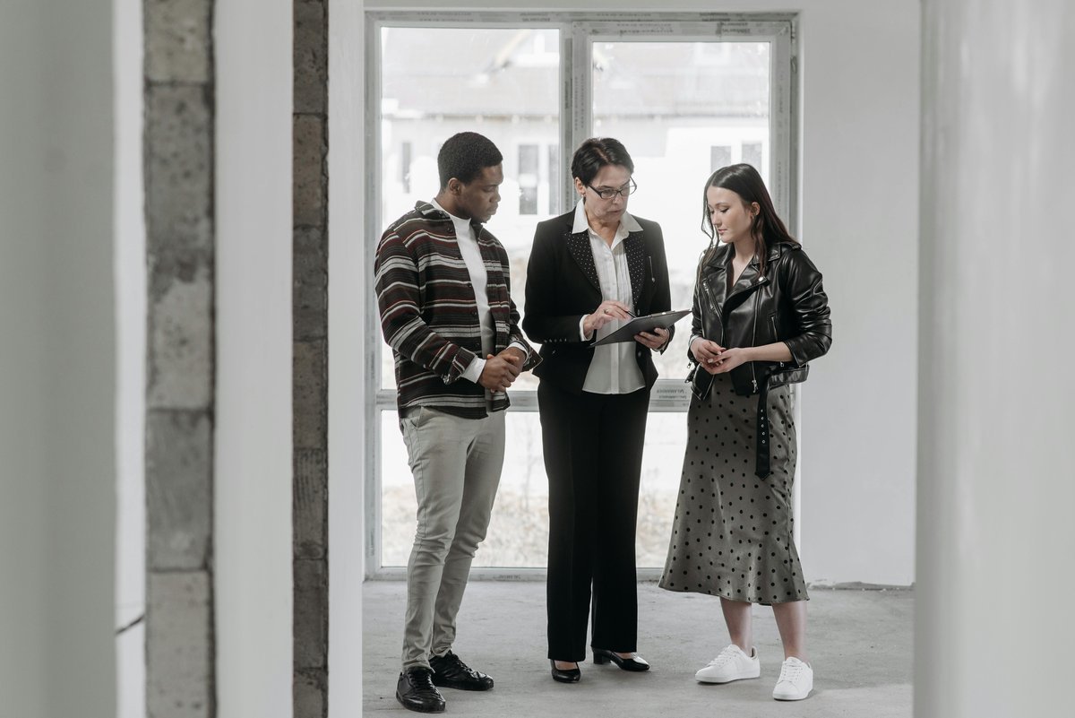 Three people standing in an empty room by a window looking at a tablet held by a woman in glasses